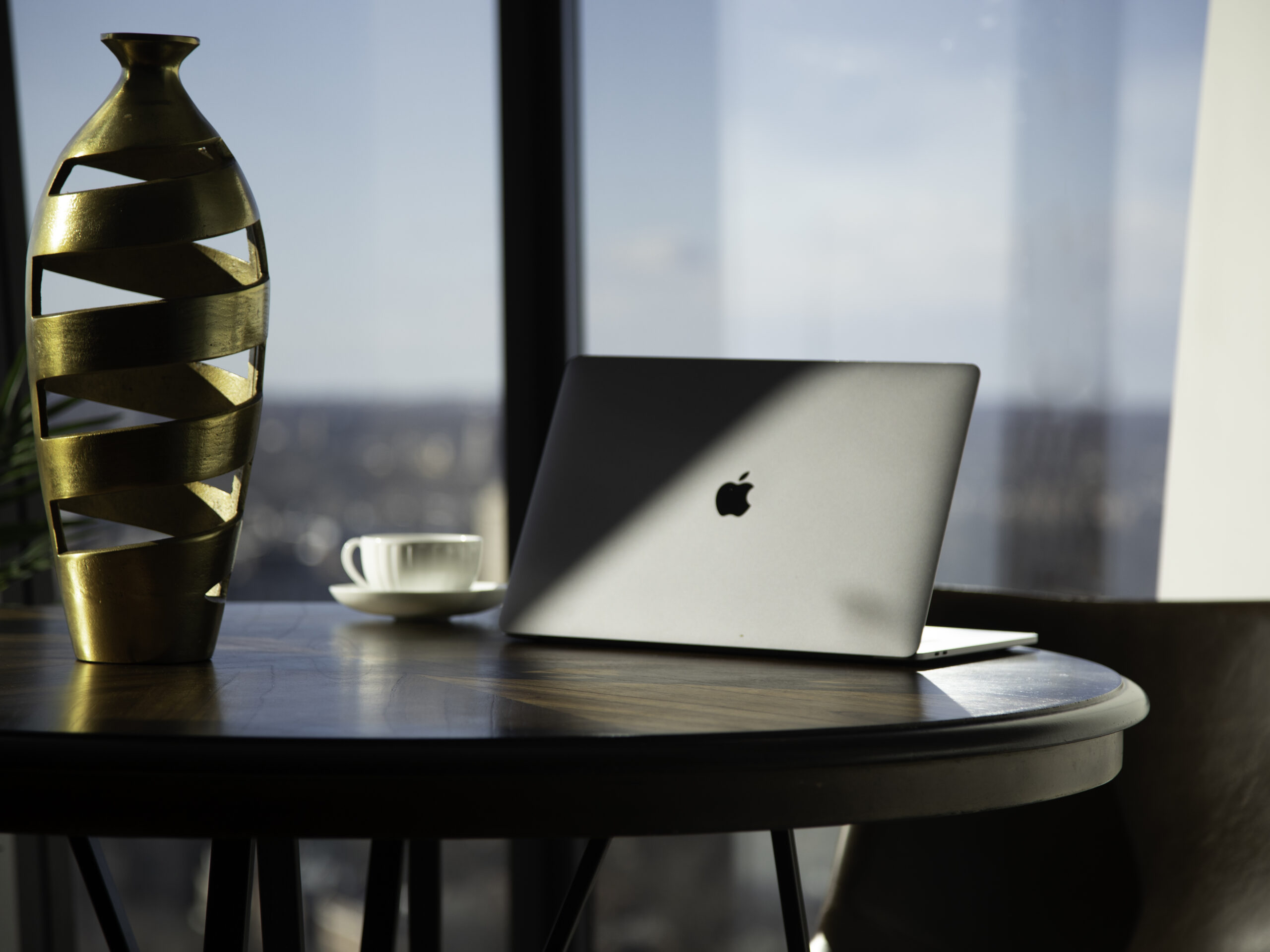 an apple laptop sits on a table next to a decorative gold vase. sunlight makes a beam across the back of the laptop. a coffee cup sits next to the laptop on the table.
