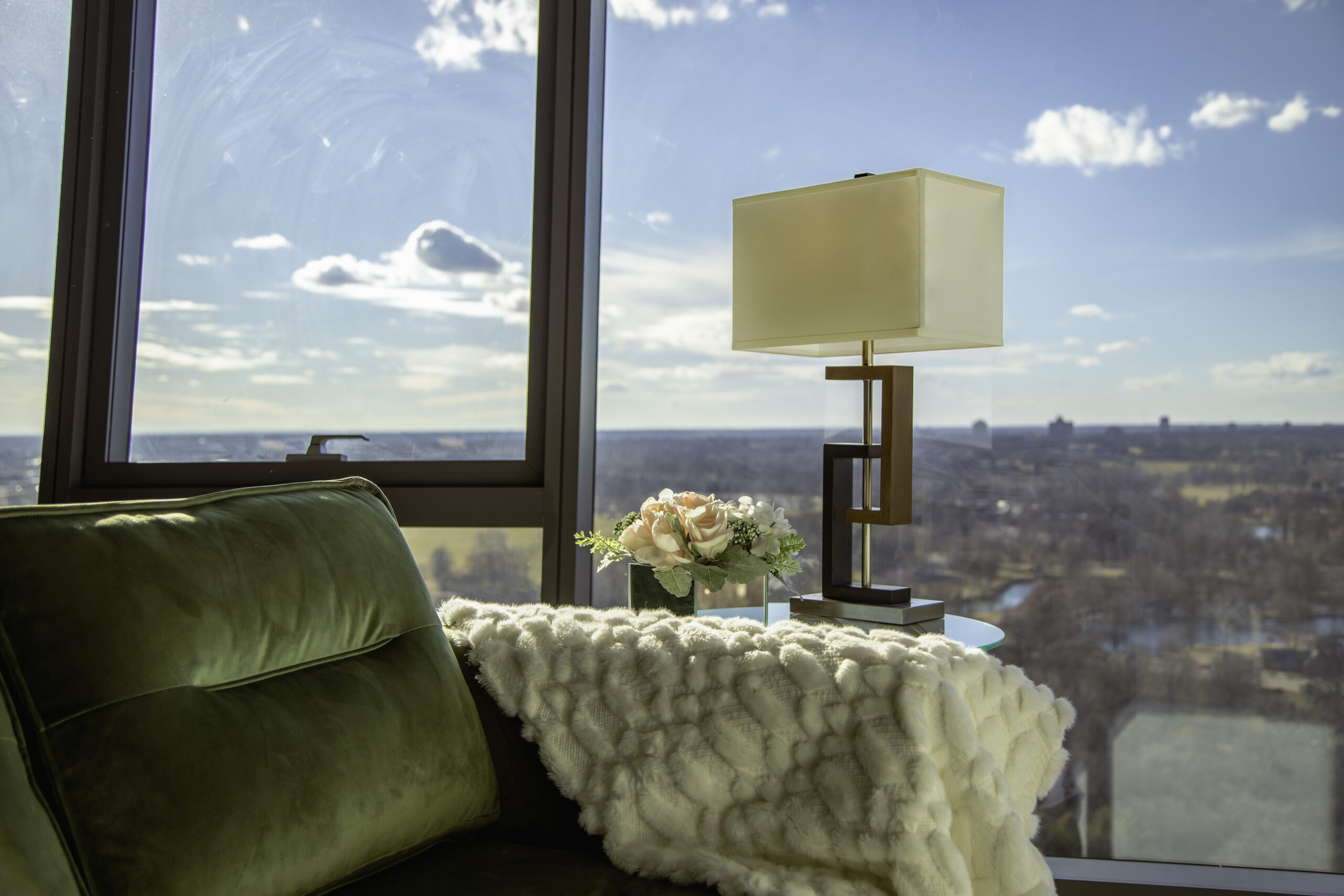 a corner of a green velvet couch has a fluffy white blanket thrown over the arm of the couch. floor to ceiling windows are visible in the background, showing a view of a city scape with a bright blue sky.